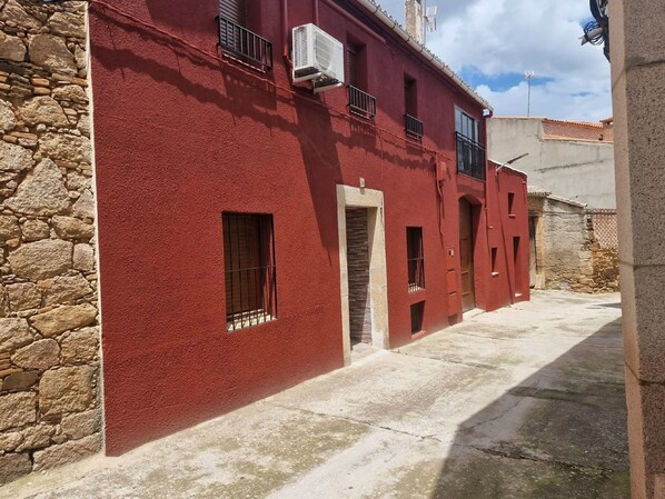 Une ruelle étroite bordée de bâtiments rouges et de murs de pierre, sous un ciel dégagé.