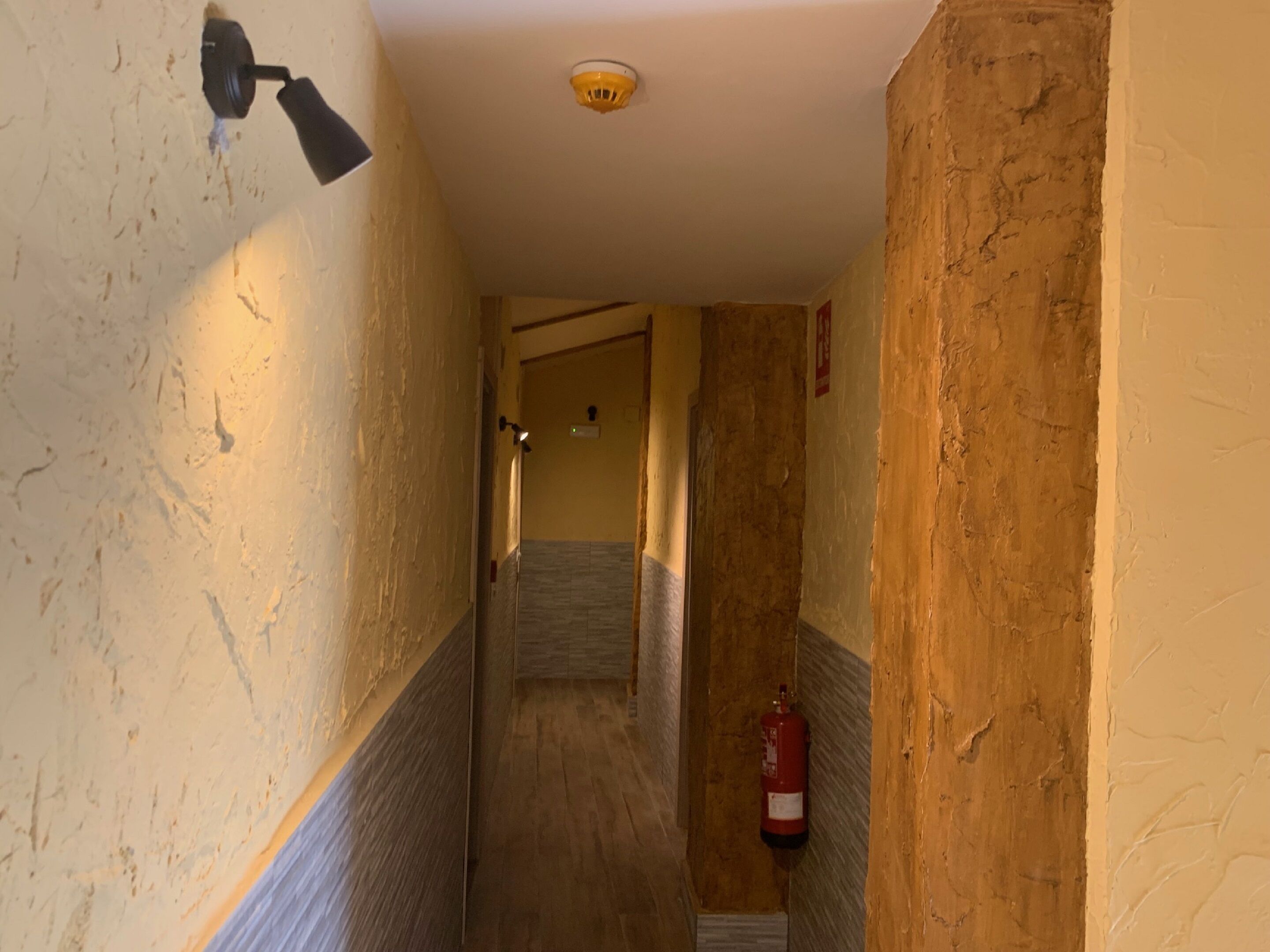 A narrow hallway with textured walls, a fire extinguisher mounted on the right, and a ceiling light fixture.