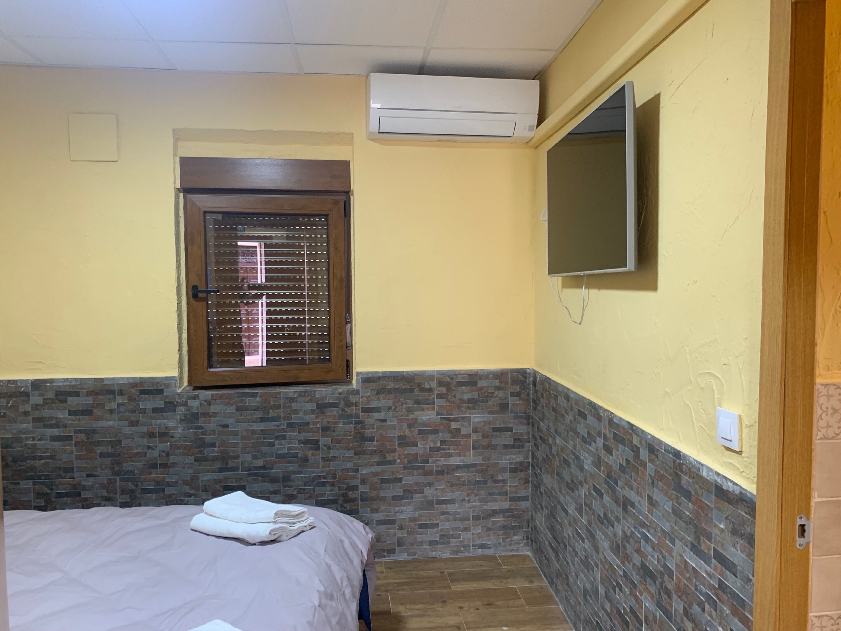 A bedroom with a bed, a window with blinds, an air conditioning unit, and a television mounted on the wall.
