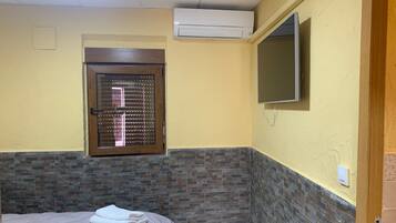 A bedroom with a bed, a window with blinds, an air conditioning unit, and a television mounted on the wall.