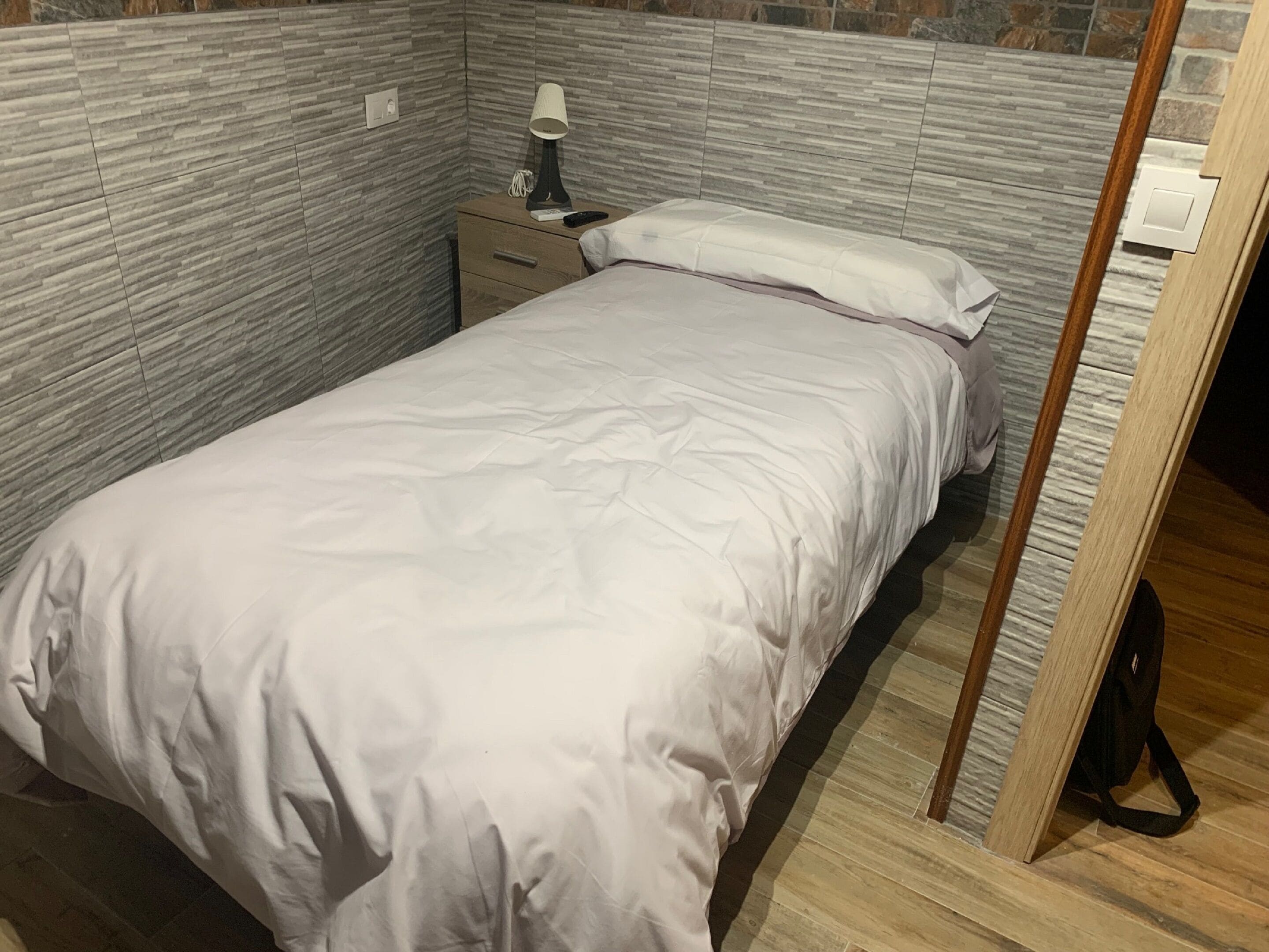 A single bed with a white duvet in a room with grey stone-patterned walls and a wooden floor.