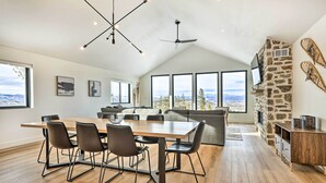 A spacious dining area with a long wooden table and modern chairs, large windows offering a view of the mountains, a stone accent wall, and a ceiling fan. - New Modern Mountain Retreat with Level 2 EV Charger (Granby)