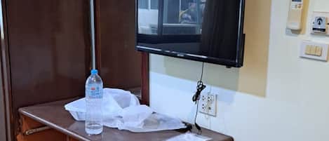 A room with a wooden desk, a television mounted on the wall, a water bottle, and a bucket on the floor.