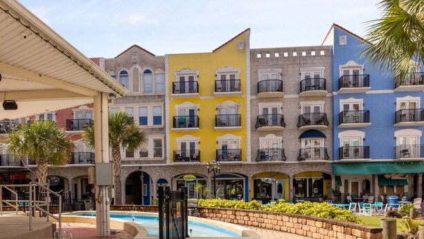 Un espace piscine agrémenté d’une fontaine, entouré d’un chemin pavé en briques. Au fond, on aperçoit des immeubles colorés à plusieurs étages, dotés de balcons.