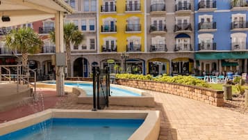 A pool area with a water fountain, surrounded by a brick walkway. In the background, there are colorful multi-story buildings with balconies.