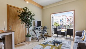 Un salon moderne avec une porte en bois, un bureau avec un ordinateur, une grande plante en pot, un canapé et une vue sur un balcon aménagé avec des sièges d’extérieur.