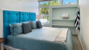 A bedroom with a blue upholstered headboard, a bed with a light blue bedspread and pillows, a bedside table, a window with a view of palm trees, and a built-in shelf with towels.