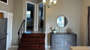 A hallway with a wooden staircase, a gray cabinet with drawers, a round mirror, and a chandelier. - Newly Renovated 2 Bedroom House (Seaside Heights)