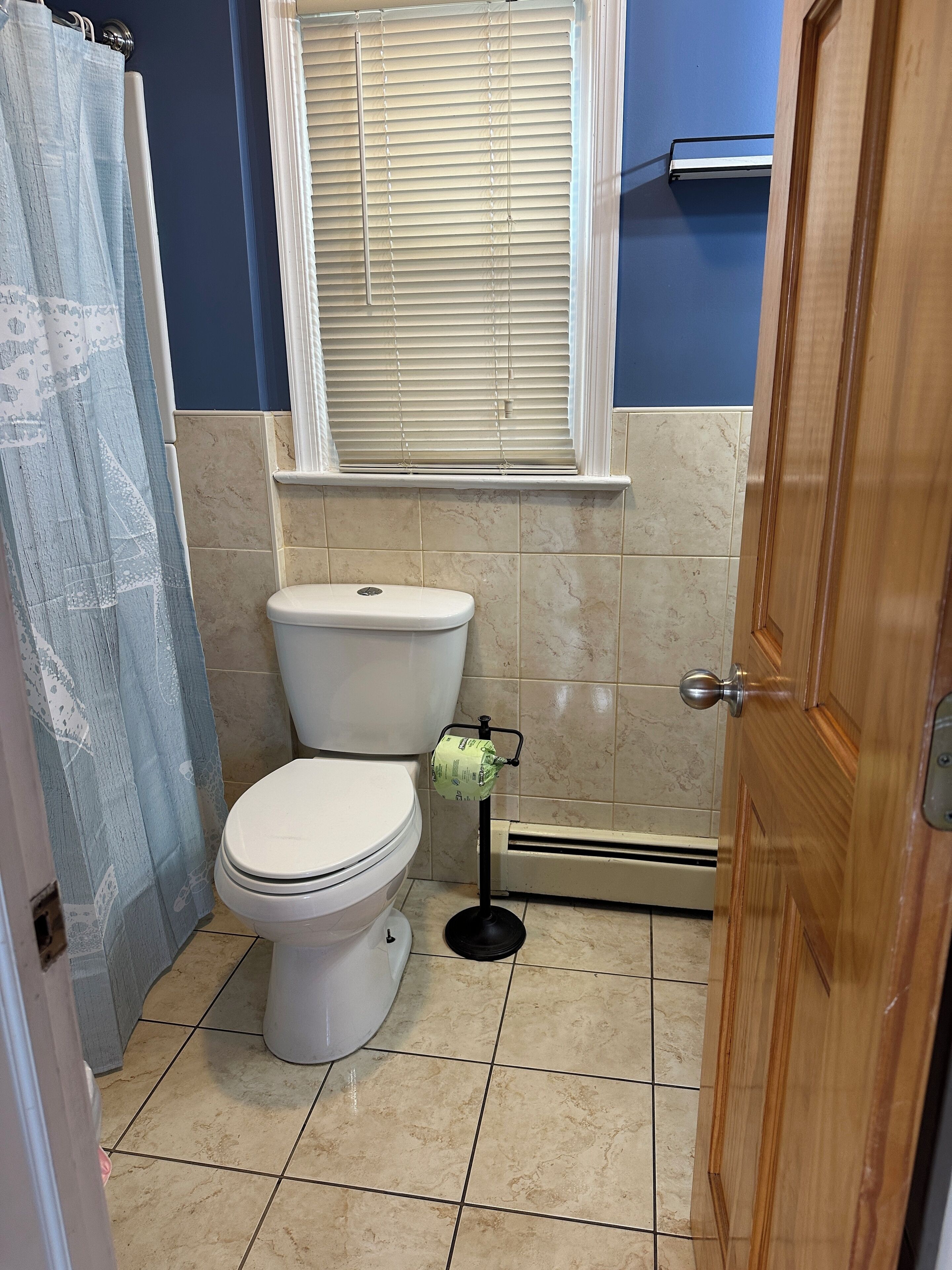 A bathroom with a toilet, a shower curtain, a window with blinds, and a door.