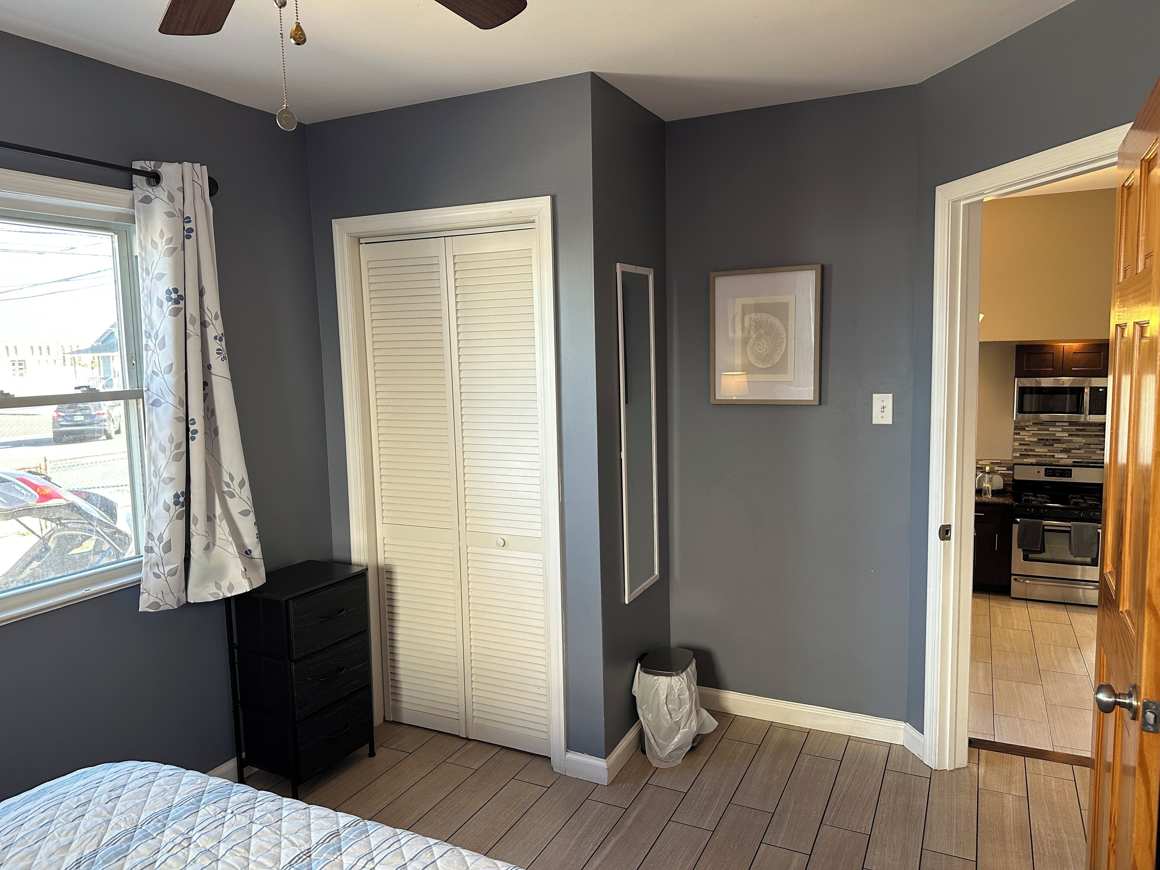 Une chambre à coucher comprenant un lit, une armoire, une fenêtre avec des rideaux, une porte donnant sur la cuisine et une petite poubelle.