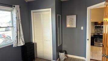 Une chambre à coucher comprenant un lit, une armoire, une fenêtre avec des rideaux, une porte donnant sur la cuisine et une petite poubelle.
