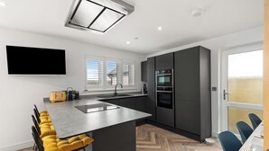 A modern kitchen with a central island, a built-in oven, and a large window with blinds. - The Paddock - Lovely Cottage with Hot Tub (Penally)