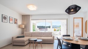 A modern living room with a sofa, two small tables, a dining table with chairs, and a large window offering a view of the outdoors. - Jardines - Acoran 2.5 RESERVE NATURE VIEW 1B (Arona)