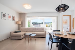 A modern living room with a sofa, two small tables, a dining table with chairs, and a large window offering a view of the outdoors.