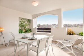 A modern dining area with a white table and chairs, a view of a desert landscape through a window, and a wall-mounted plant.