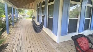 Une véranda couverte avec un plancher en bois, un hamac et un fauteuil en osier avec un coussin rouge.