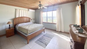 Une chambre avec un plafond en bois, un lit avec une tête de lit, une table de chevet, une commode, une fenêtre avec des rideaux et un ventilateur de plafond.