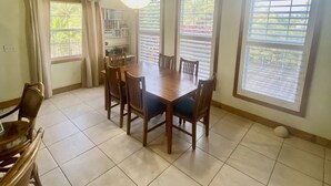 Un coin repas avec une table et des chaises en bois, de grandes fenêtres équipées de stores et un ventilateur de plafond.