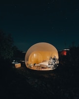 Estrutura esférica iluminada com uma cama, mesa e banheira, cercada por árvores e céu noturno.