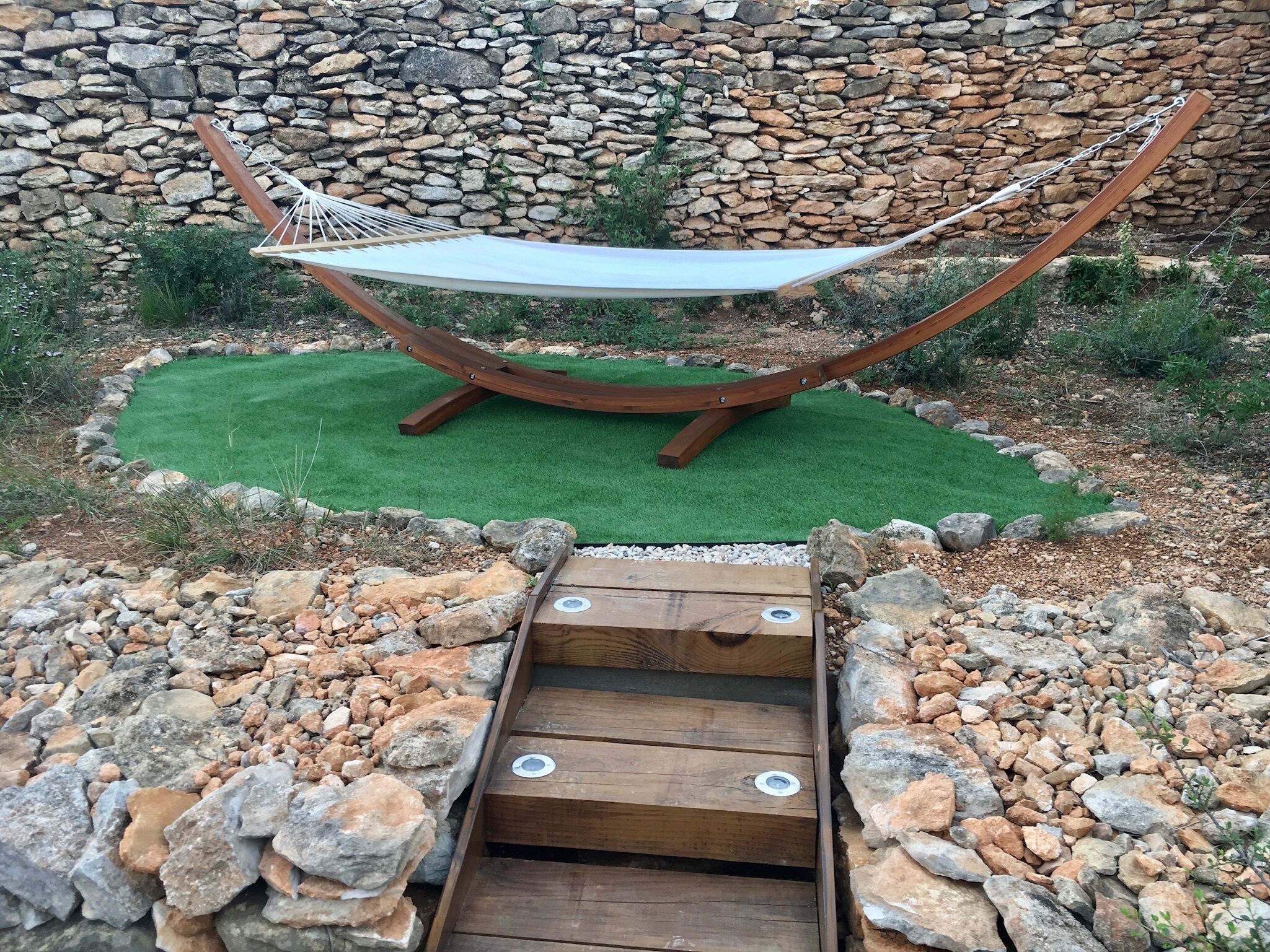 A hammock suspended between two wooden posts, set against a stone wall and surrounded by a rock garden.