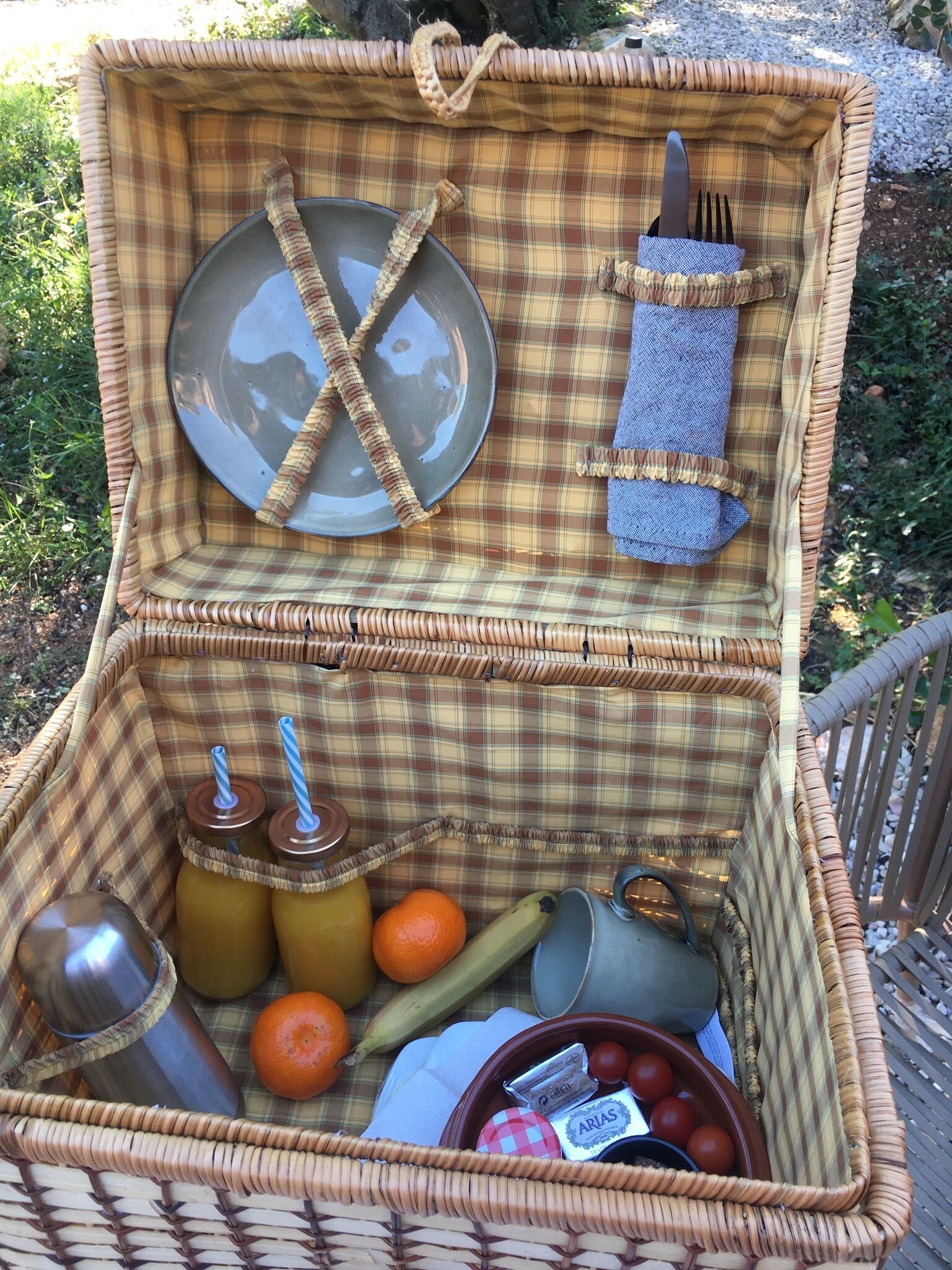 A wicker picnic basket with a plaid interior, containing a metal plate, a fork, a knife, a bottle, a thermos, a banana, an orange, a bottle of water, a bowl of tomatoes, and a small container of cheese.