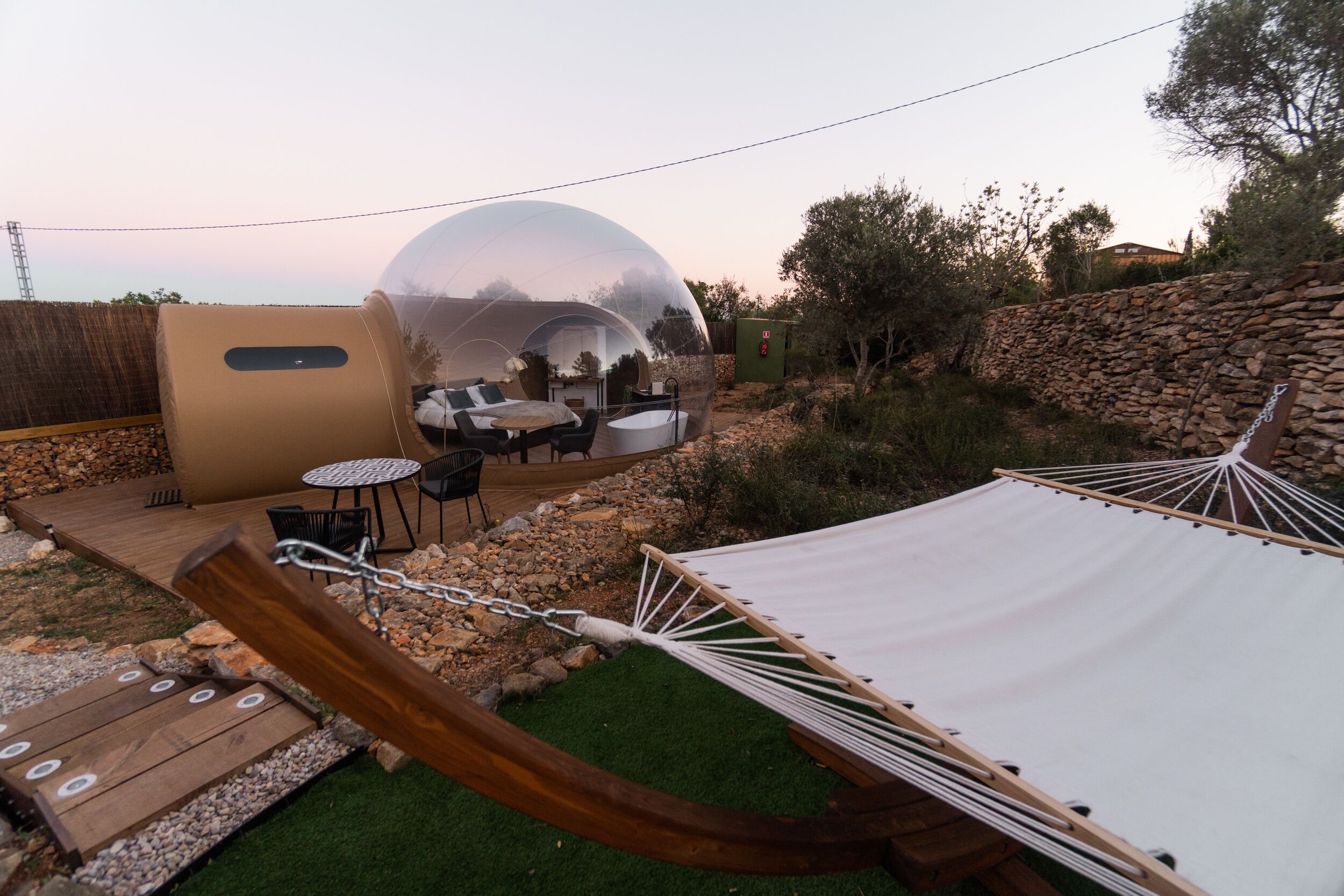 A transparent dome structure with a seating area inside, surrounded by a stone wall and greenery.