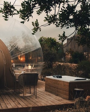 Une structure en forme de dôme transparent, abritant une table et des chaises, entourée d’une terrasse en bois et d’un mur de pierre.