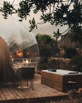 Estrutura em cúpula transparente com uma mesa e cadeiras no interior, rodeada por um deck de madeira e uma parede de pedra.