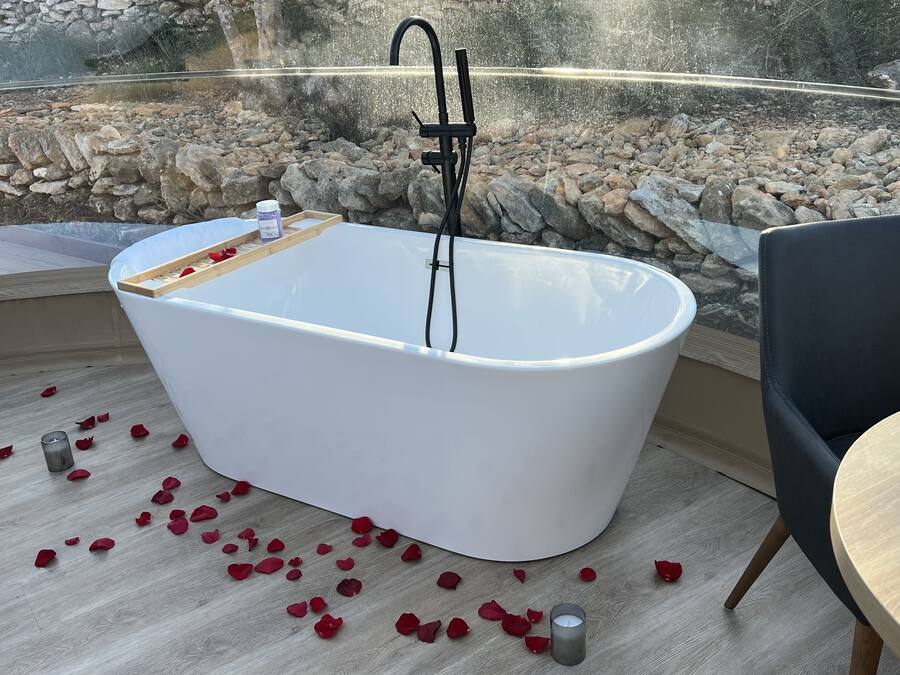 Un baño moderno con una bañera independiente, una bandeja de madera con pétalos de rosa y una mesa de cristal con una copa de vino.