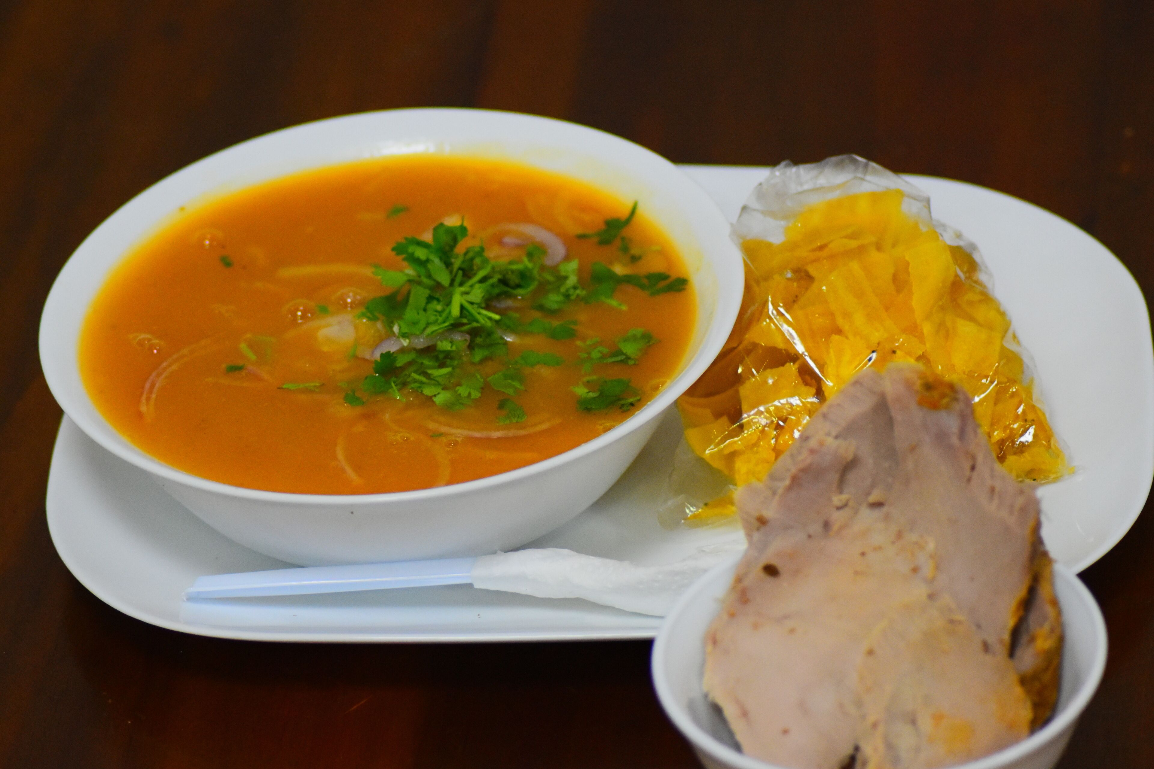 A bowl of soup with a slice of meat and a piece of bread on a white plate.