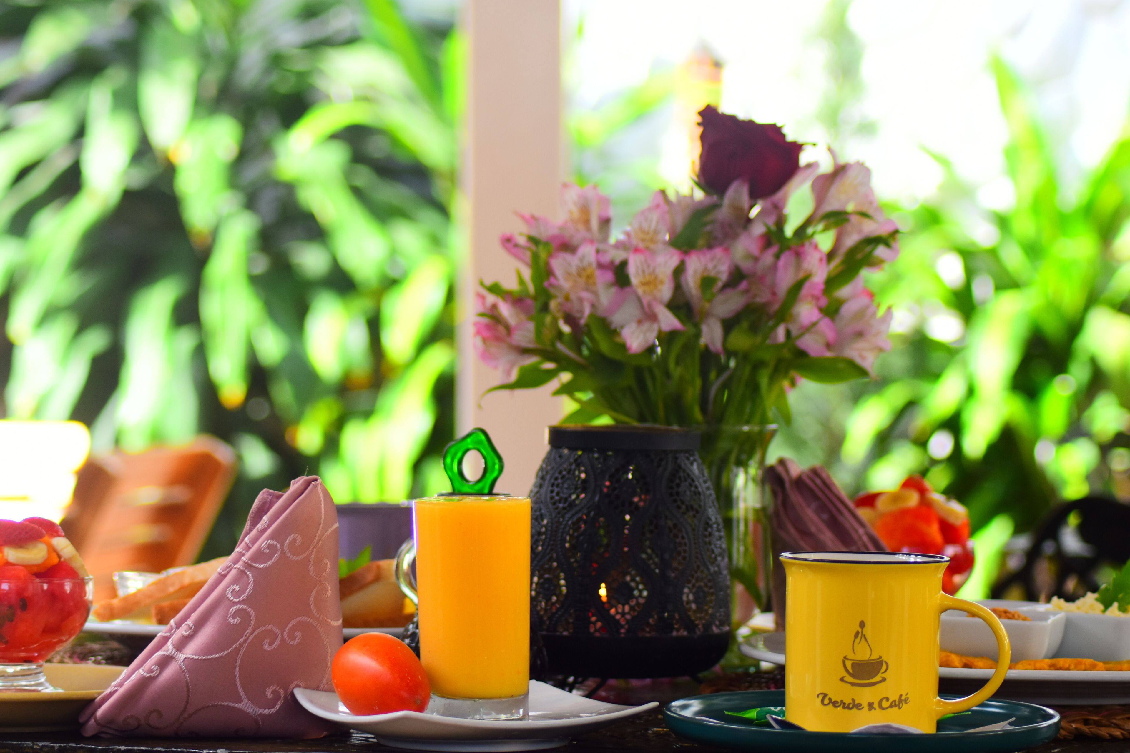 A breakfast table with a glass of orange juice, a yellow mug, a vase of flowers, and a plate with a red tomato.