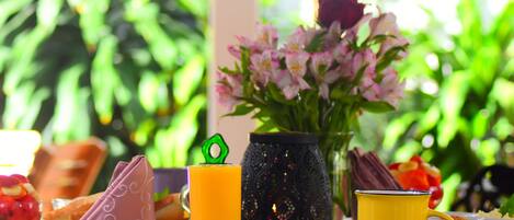 Una mesa de desayuno con un vaso de zumo de naranja, una taza amarilla, un jarrón de flores y un plato con un tomate rojo.