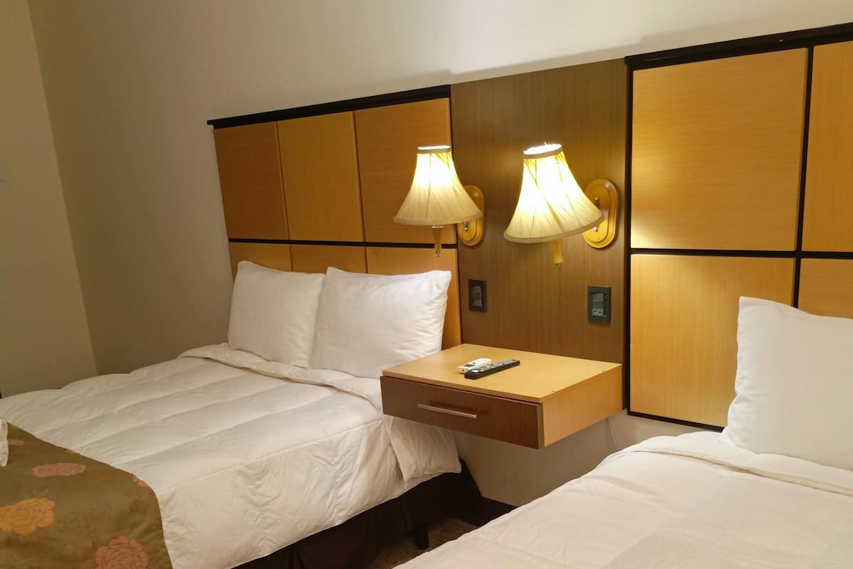 A hotel room with two beds, each with white bedding and floral-patterned covers. There are two bedside tables with lamps, and a small nightstand with a remote control.
