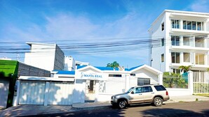 Un hotel moderno con entrada protegida por una reja, un SUV estacionado y un cielo azul despejado.