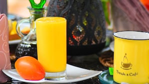 Una mesa dispuesta con un vaso de jugo de naranja, un plato con un tomate, una botella verde y una taza amarilla con diseño de taza de café.