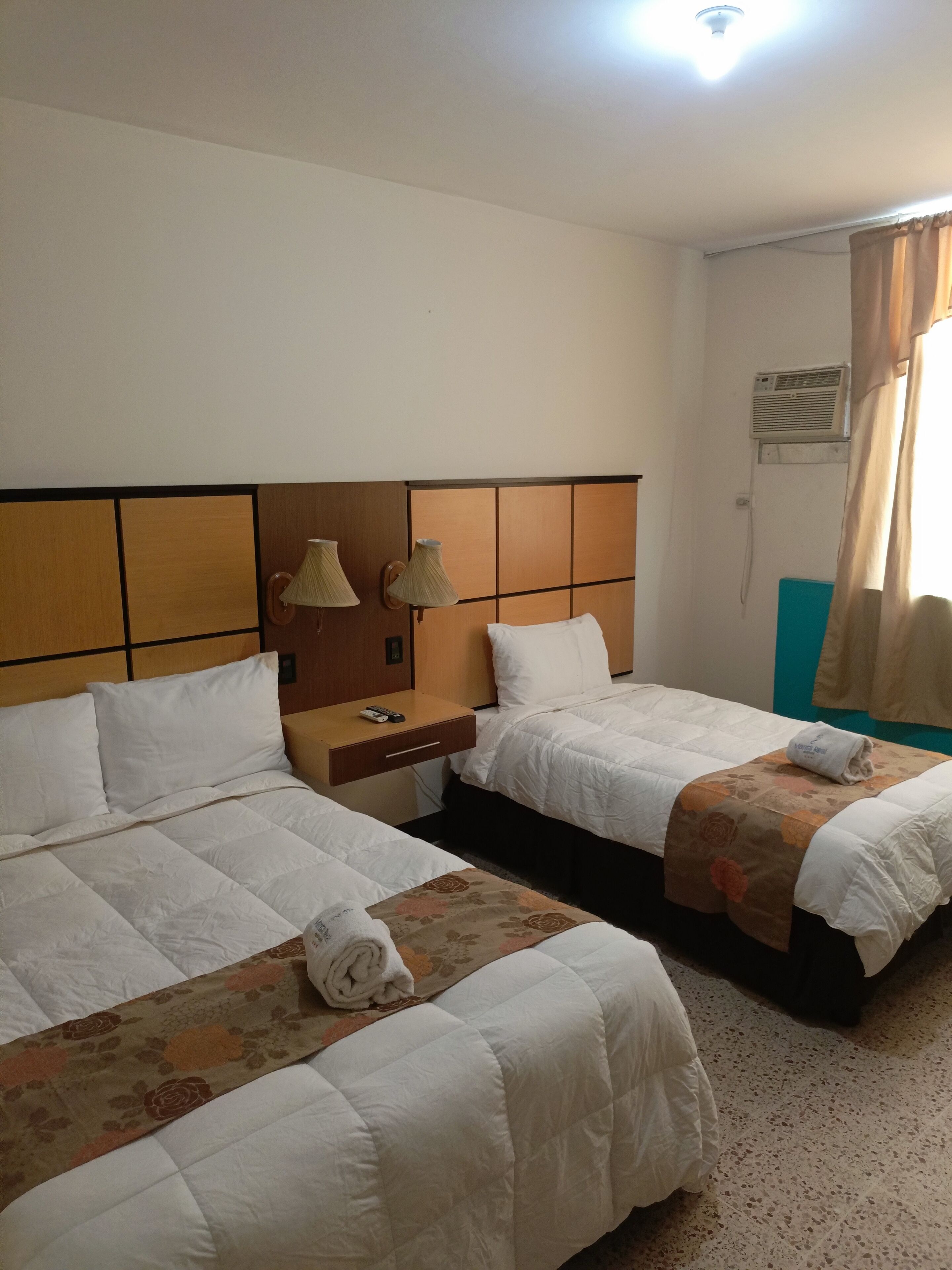 A hotel room with two beds, a nightstand, a lamp, an air conditioning unit, and a window with curtains.