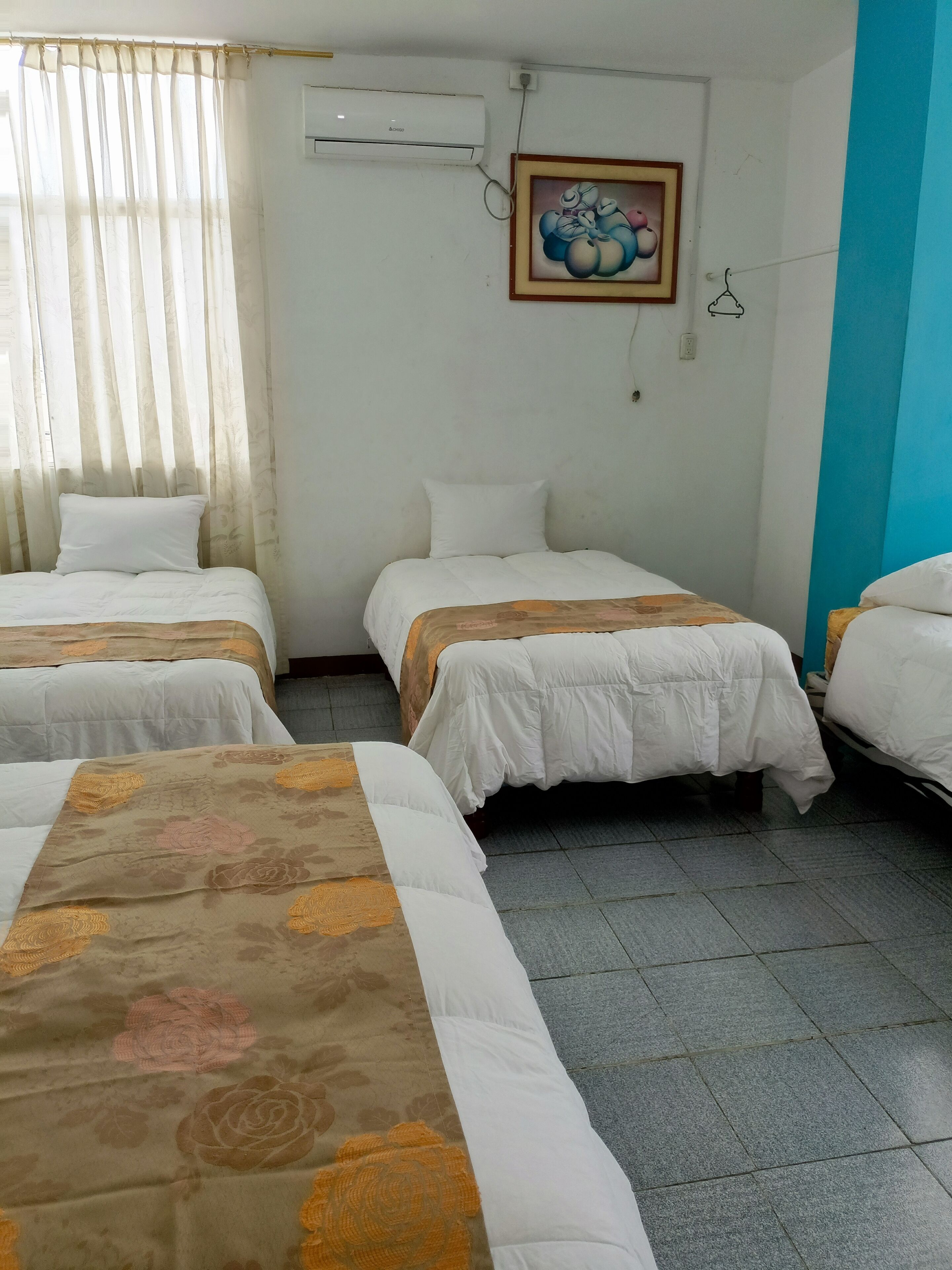 Une chambre d’hôtel avec trois lits, une fenêtre avec des rideaux légers, un appareil de climatisation et un tableau encadré au mur.