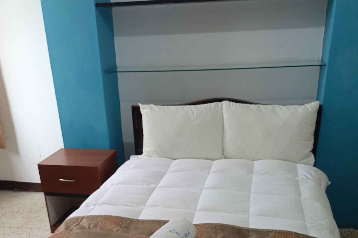 A neatly made bed with white and brown bedding, a folded towel, and a wooden nightstand with a drawer.