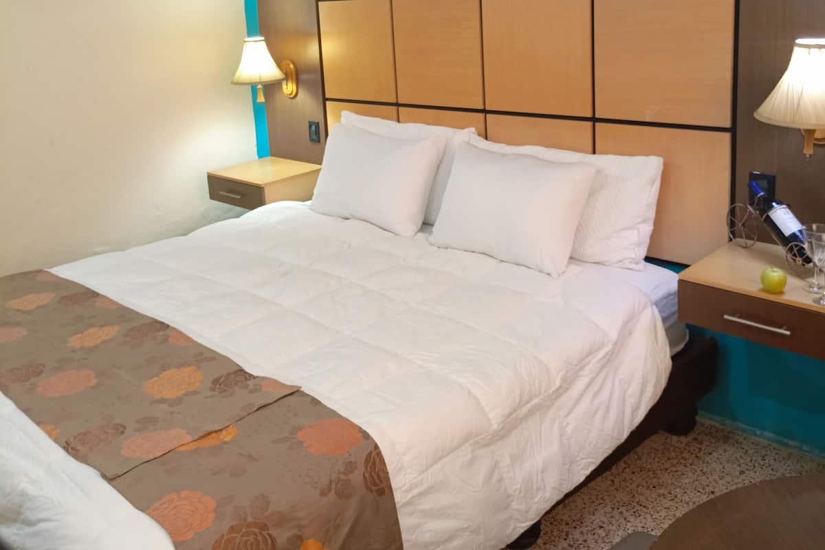 A hotel room with a large bed, a wooden headboard, two bedside tables with lamps, and a small round table with a bottle and glass.