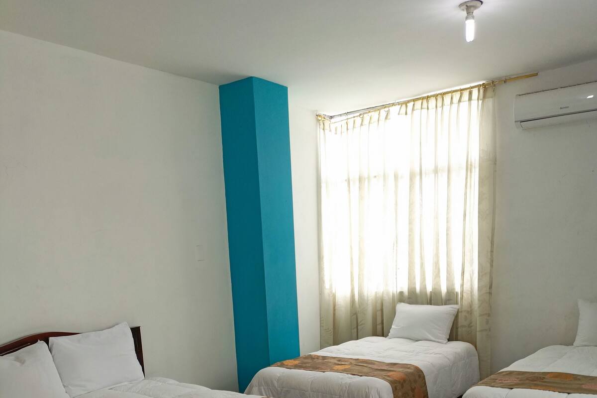 A hotel room with three single beds, a blue accent wall, a window with sheer curtains, and an air conditioning unit.