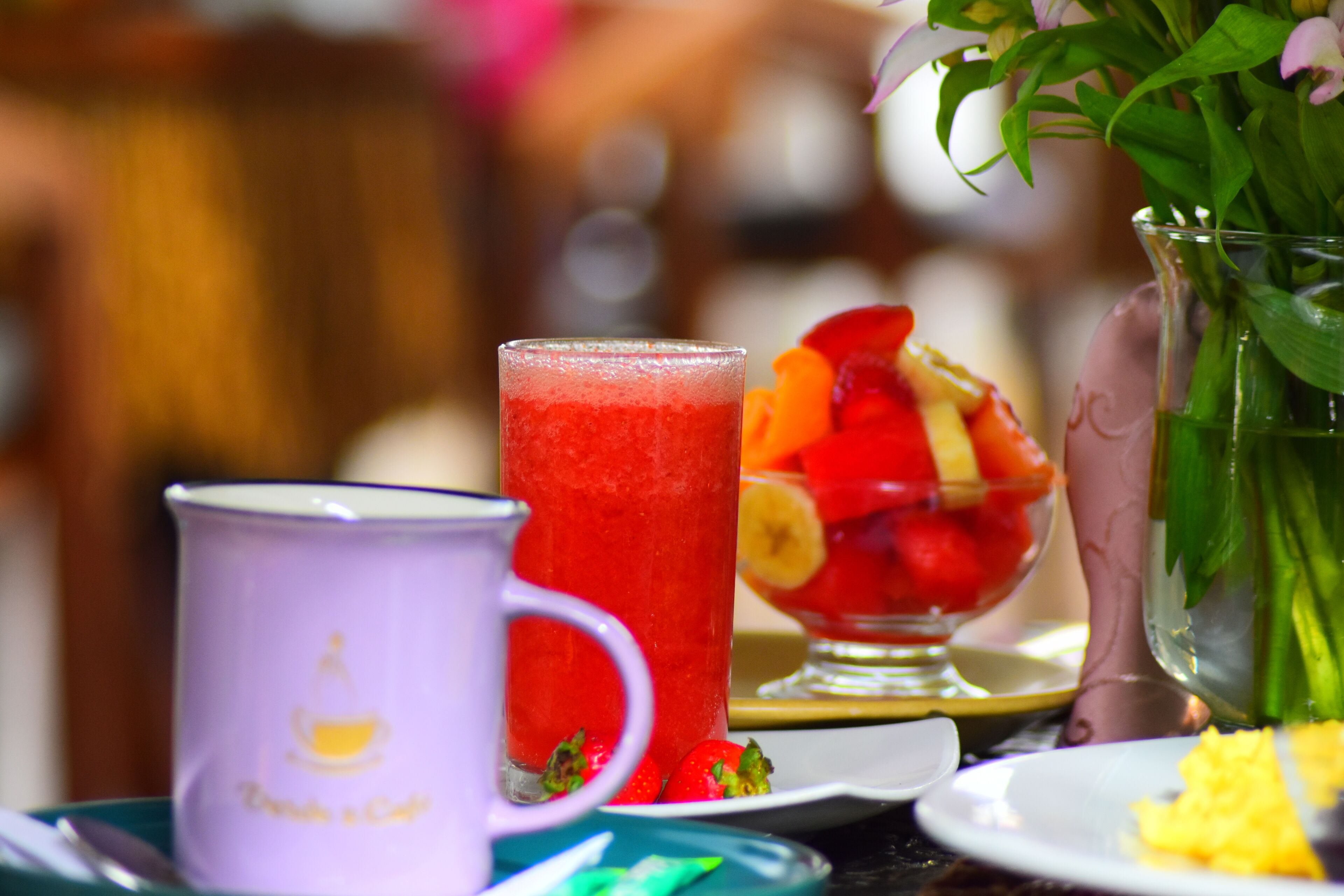 A table with a purple mug, a tall glass with a red drink, a bowl with fruit, and a vase with flowers.