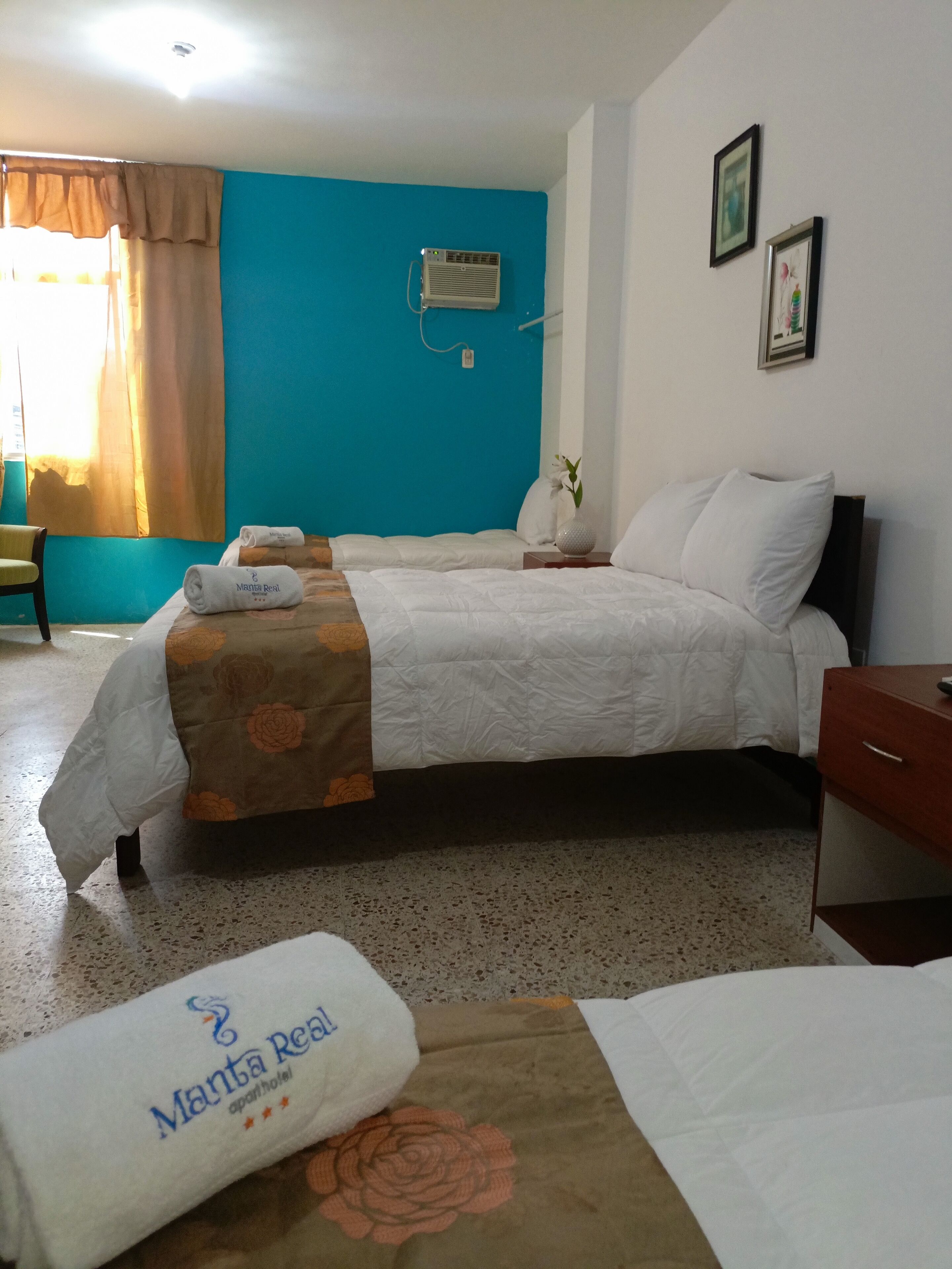 Une chambre d’hôtel avec deux lits, une fenêtre avec des rideaux, un appareil de climatisation et une petite table sur laquelle se trouve un vase.