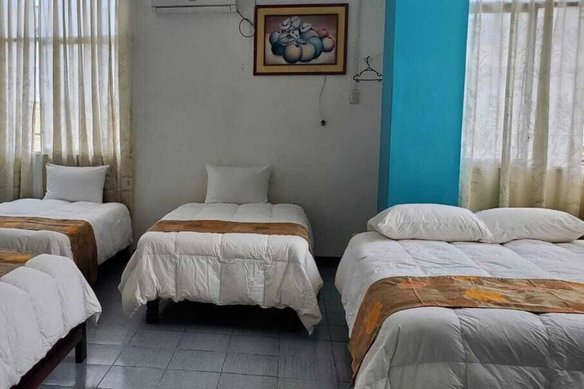 A room with four single beds arranged in a row, a painting on the wall, and a window with sheer curtains.