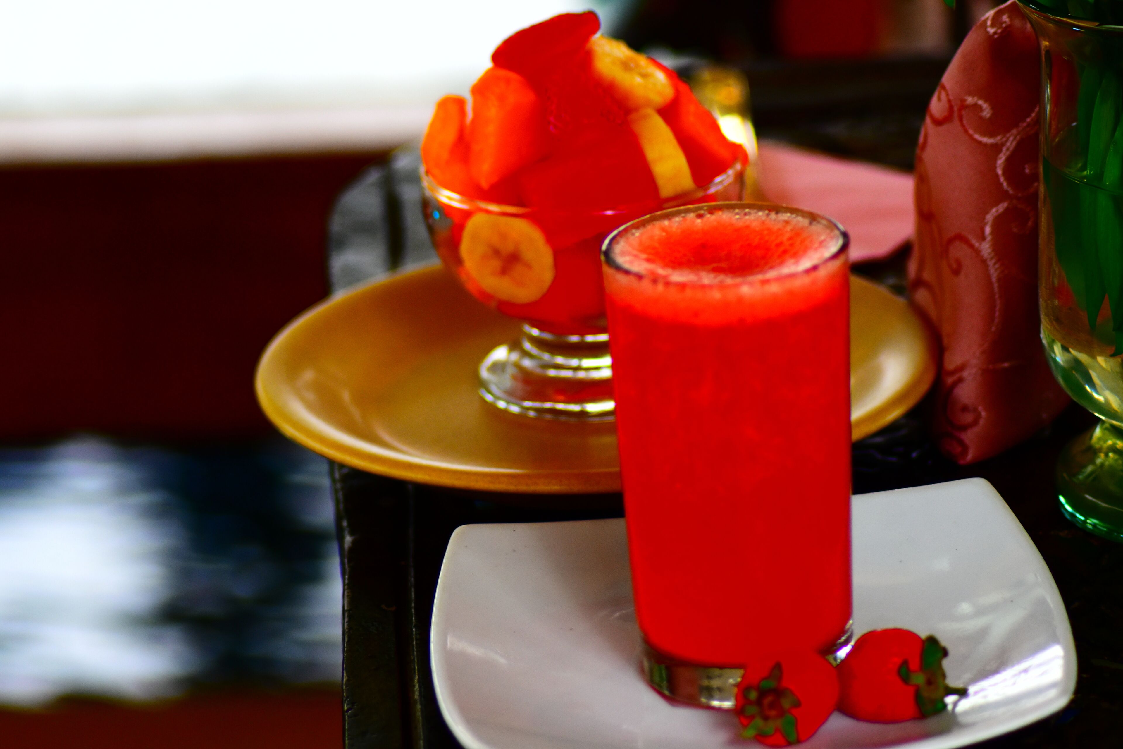 Un vaso de bebida roja sobre un plato blanco, con un cuenco pequeño de fruta al fondo.