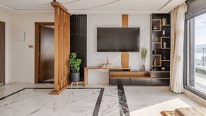 A modern living room with a large flat-screen TV, a wooden entertainment unit, a potted plant, and a large window offering a view. - Appartement Haut Standing Tanger (Tangier)