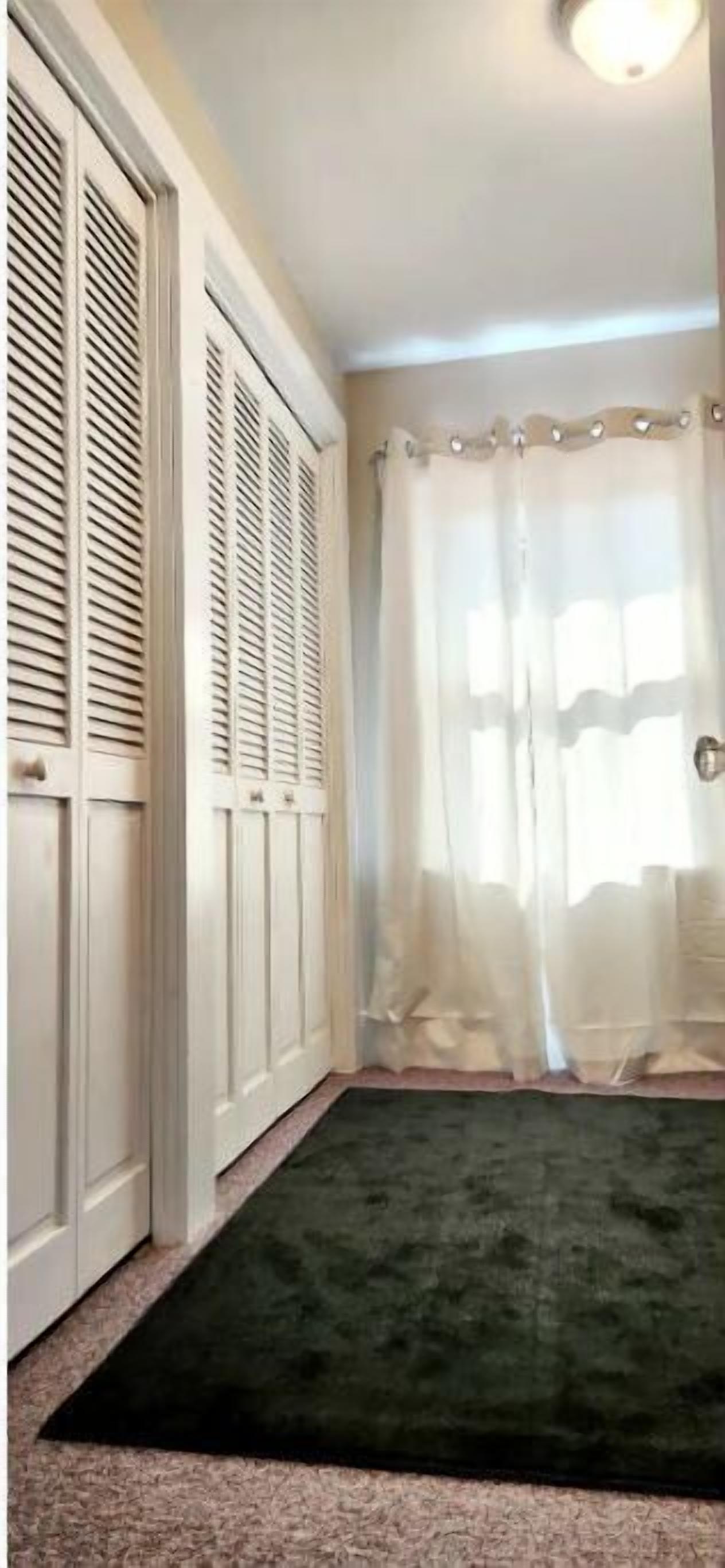 A hallway with a dark green carpet, white doors with louvered tops, and a sheer white curtain.