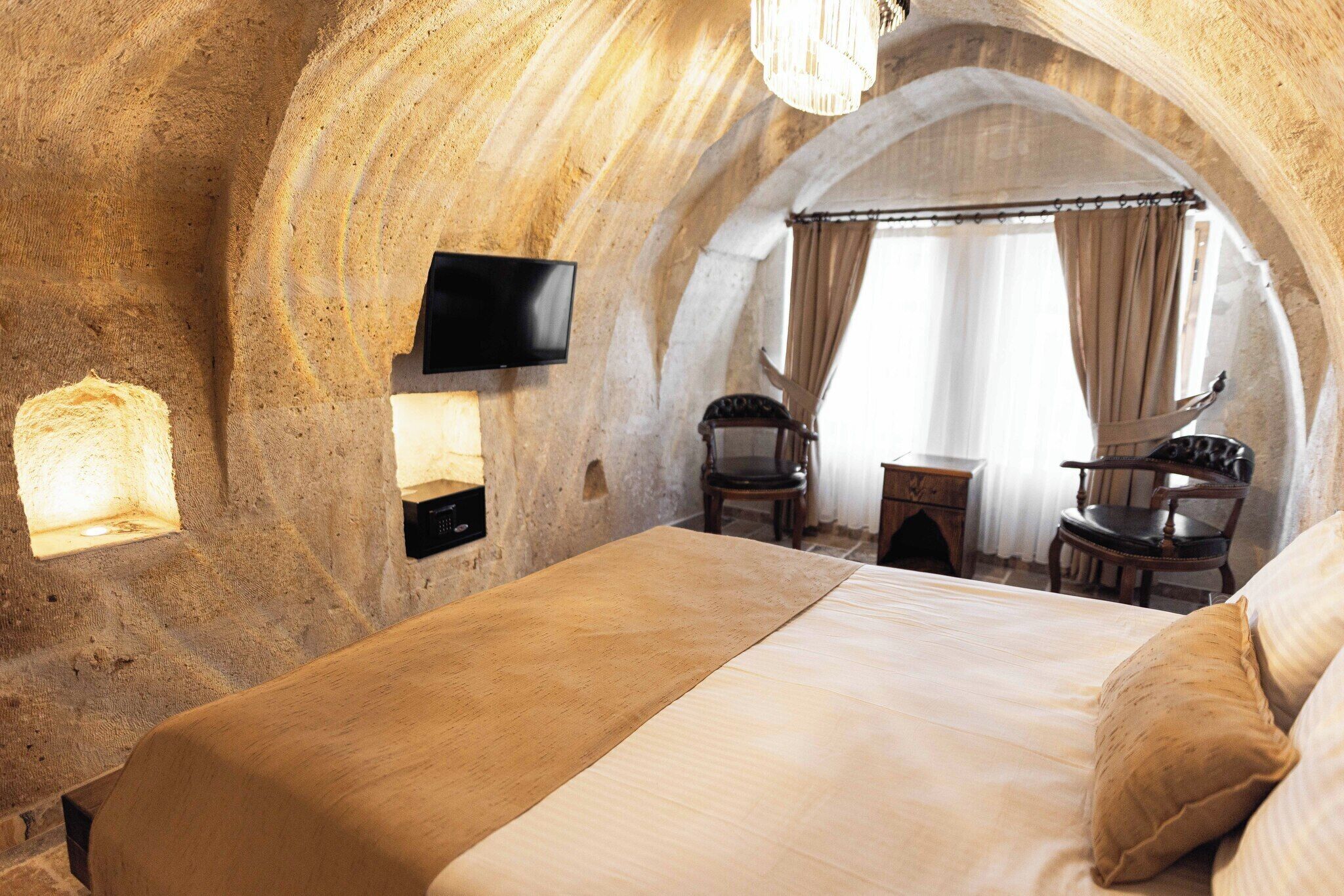 A cozy, stone-walled bedroom with a bed, a flat-screen TV, a small table, two chairs, and a window with curtains.