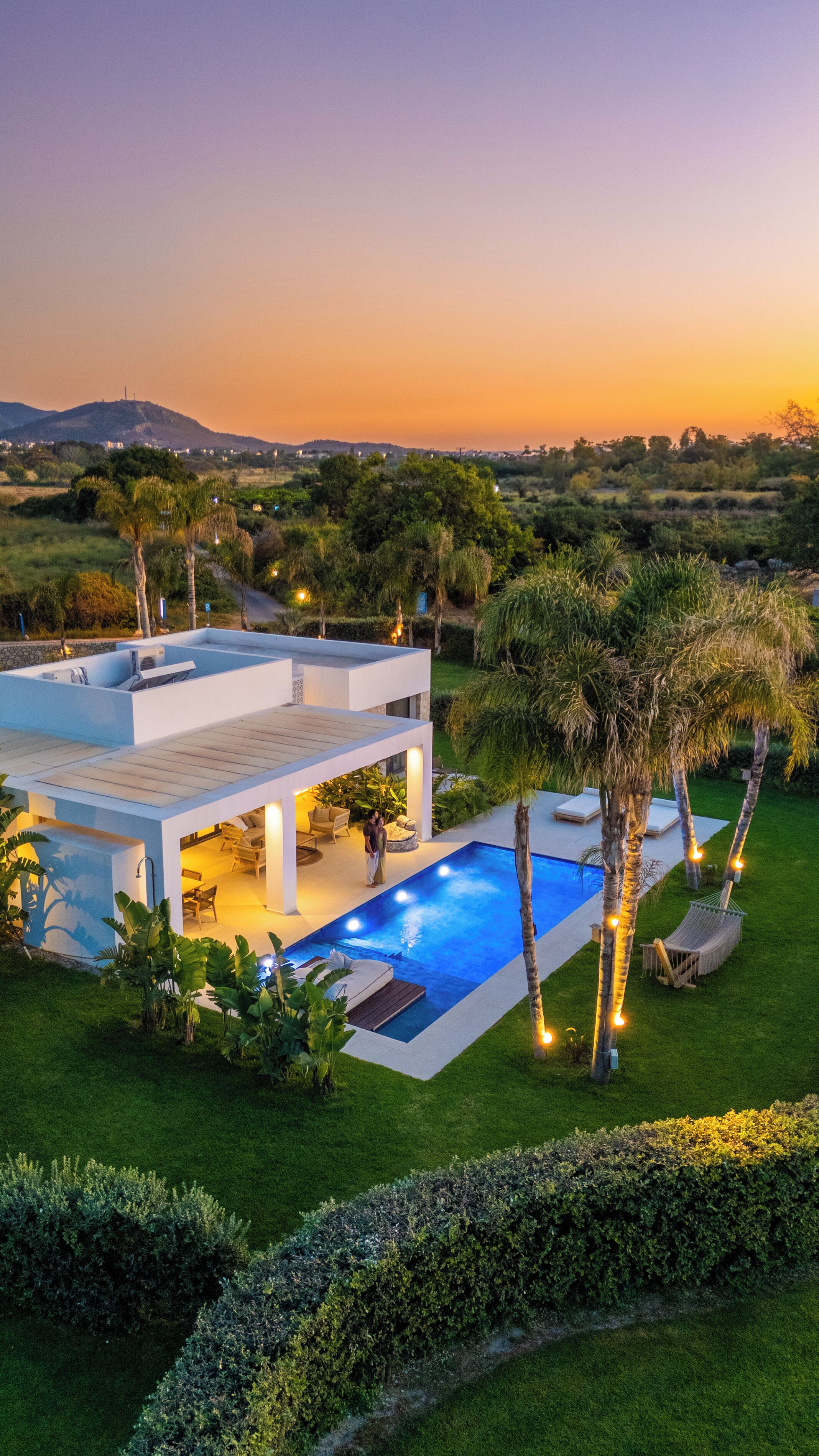 A modern house with a swimming pool, surrounded by lush greenery and a sunset in the background.