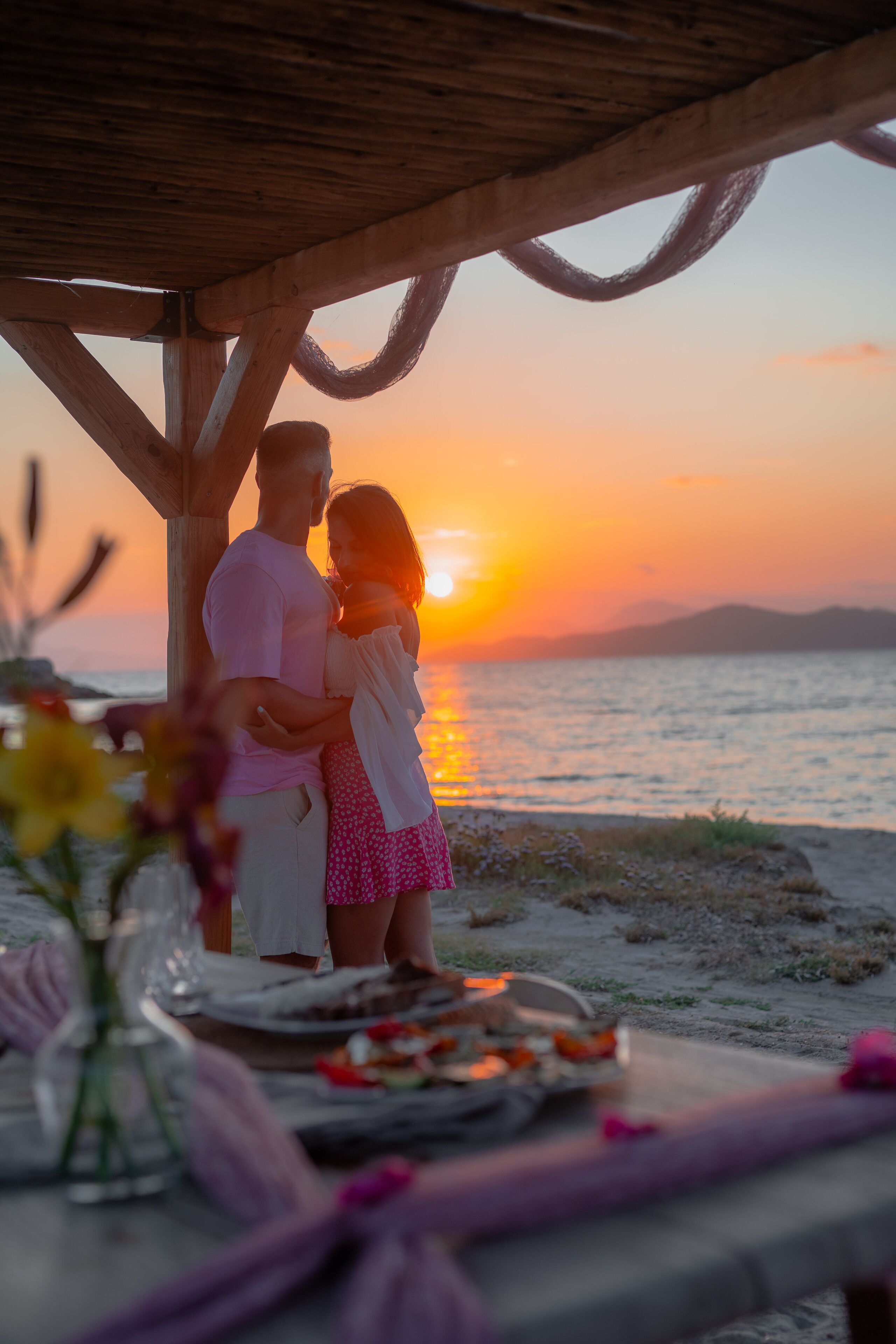A couple embracing at a wooden table with a sunset view.