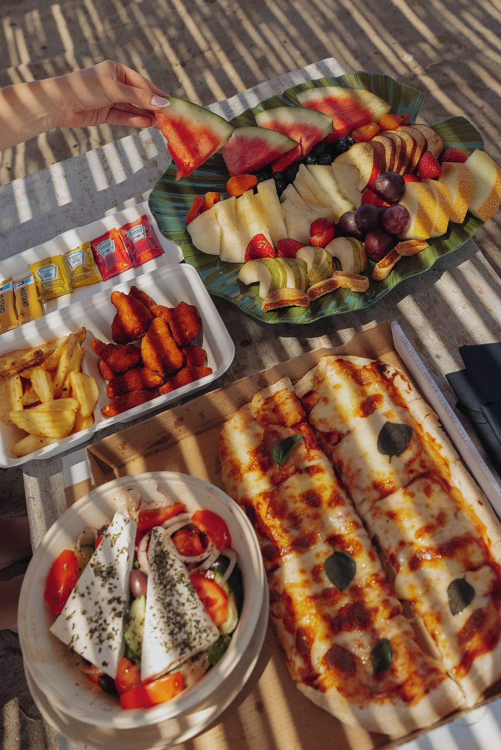 A table with various food items including a plate of sliced fruits, a tray of fries, a bowl of salad, and a pizza.